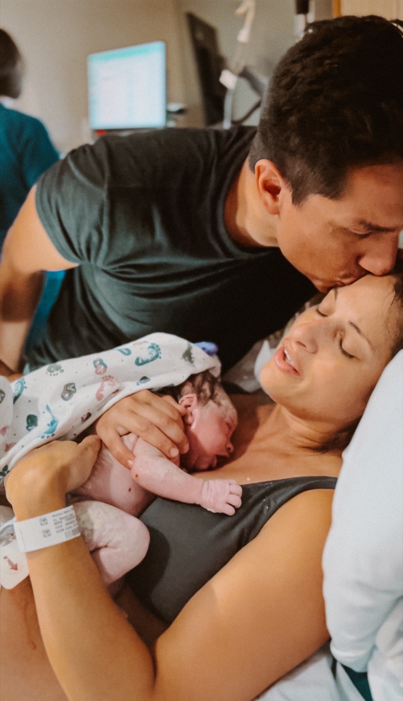 Partner kissing mother's head as she holds their newborn skin to skin